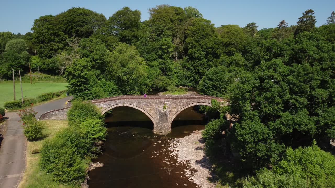 Man on a double arched bridge over a shallow river surrounded by trees