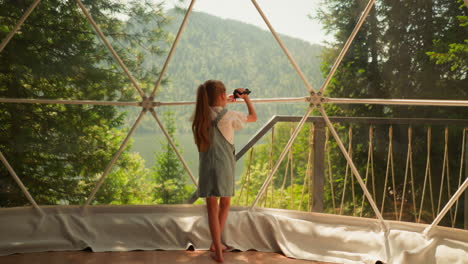 child records video with camera at glamping. little girl uses videocamera to take shot through panoramic window. kid imitates camerawoman at resort