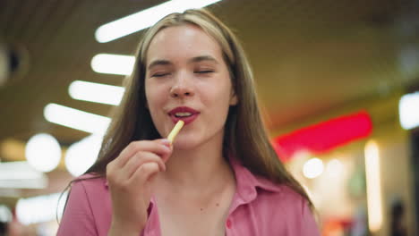 mujer sentada en un restaurante toma una patata frita, sonríe, toma un bocado, cierra los ojos de satisfacción, y sacude la cabeza, saboreando el delicioso sabor, con luces brillantes del restaurante en el fondo