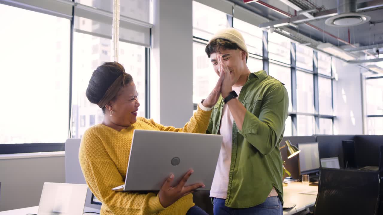 Diverse coworkers entering office, opening laptop and laughing while reviewing data at white desk