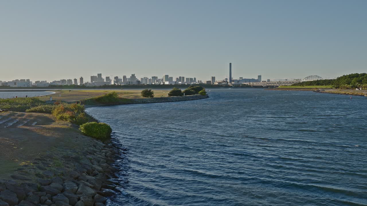 A wide shot of a river bend with a rocky bank and green parkland, leading the eye toward a distant urban skyline and tall smokestack