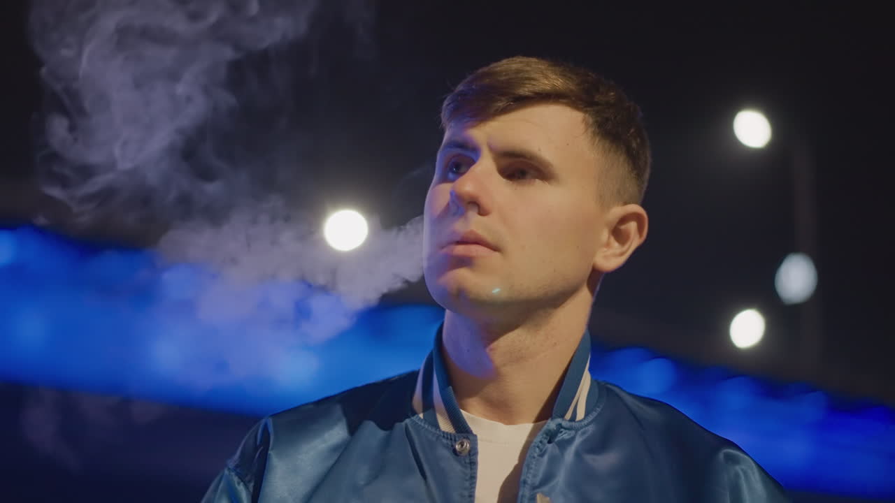 네온 불빛 다리 아래에서 한 백인 남성이 증기를 내뿜고 있어요. 짙은 연기 구름, 손에 들린 베이핑 기기, 보케 하이라이트가 있는 영화 같은 옆모습, 사색에 잠긴 시선과 분위기 있는 야간 분위기가 돋보입니다.