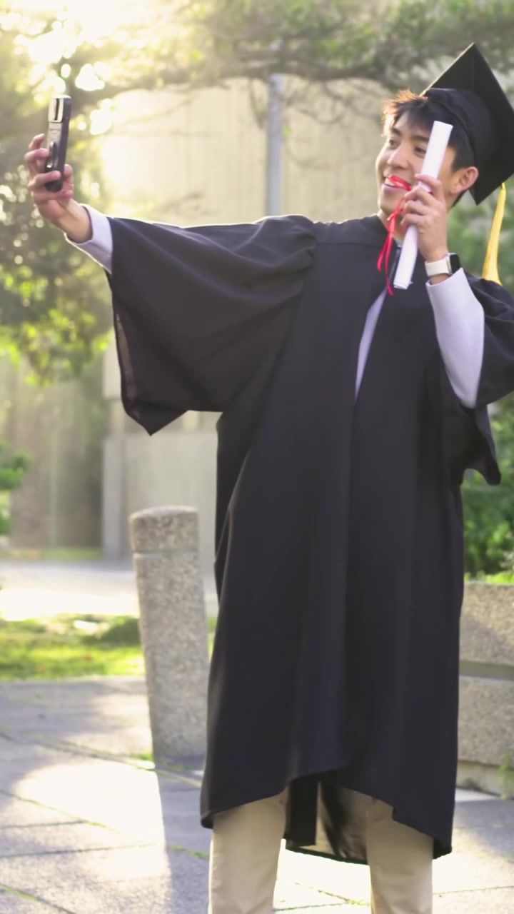 Video vertikal: Lulusan mengenakan toga dan topi wisuda memegang ijazah, menggulir ponsel di kampus sambil merayakan.
