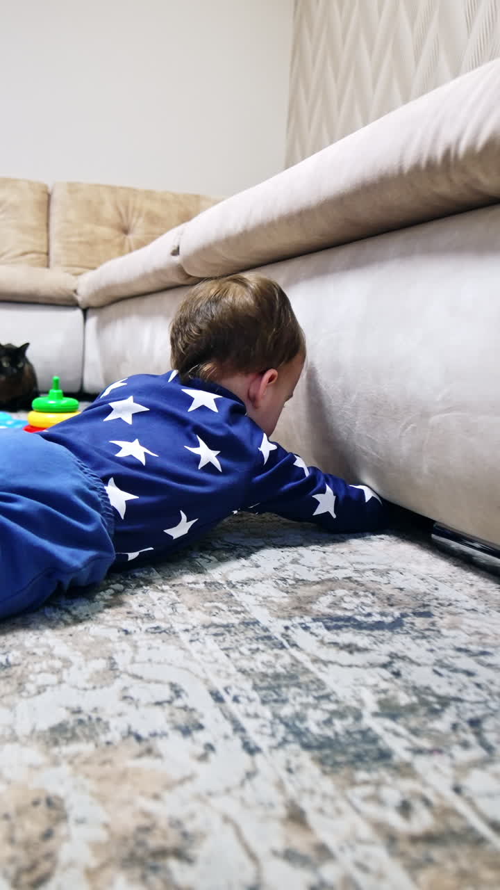 Toddler wearing blue clothes lies on the floor looking under the sofa. Baby pulls the phone from under the couch and then pushes back. Vertical video