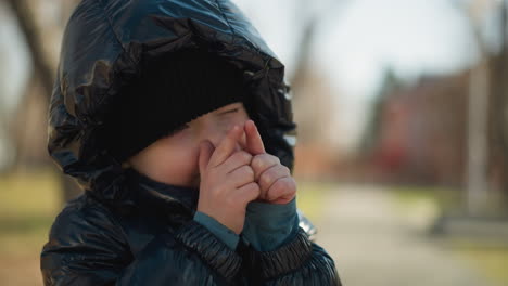 a young child playfully holding his hands close to his face, slightly closes his eyes, the child is dressed in a shiny black jacket, with an adult in a brown jacket nearby, with blurred background