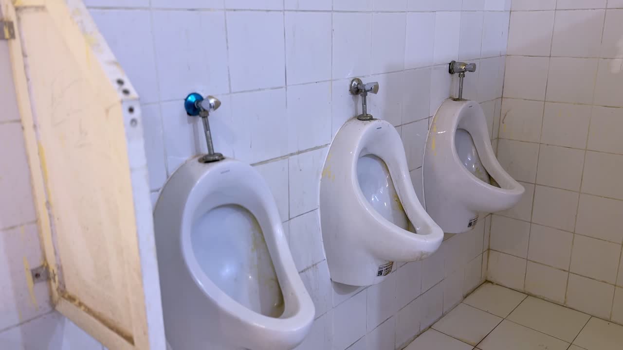 Vista laterale di tre orinatoi in ceramica a parete con macchie visibili e pannello divisorio all'interno di un bagno pubblico piastrellato e datato, che mostra le attrezzature sanitarie e l'impianto idraulico interno.
