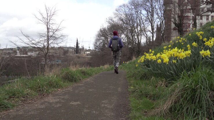 Ripresa panoramica di una ragazza che passa, a passeggio con narcisi gialli in primo piano in una giornata soleggiata e ventosa con vista sulla città di Edimburgo, Scozia, sullo sfondo.