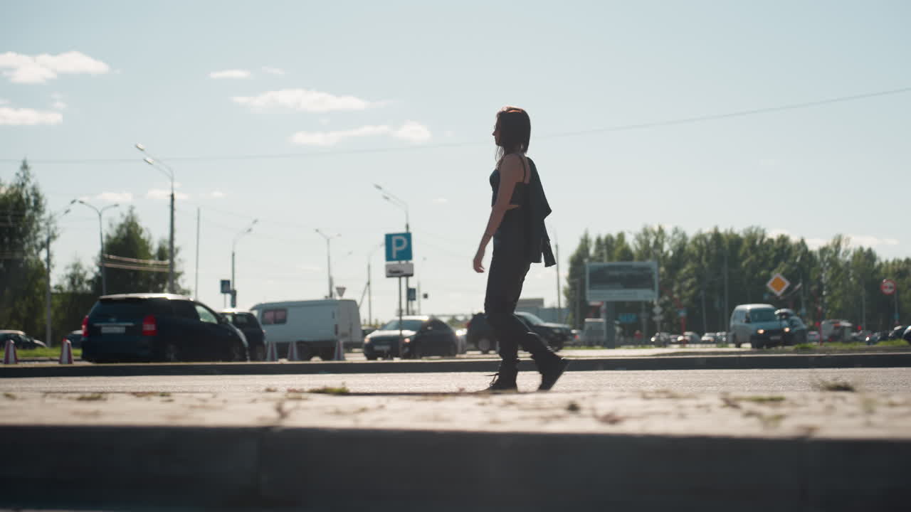 Side view of young woman in black outfit walking confidently with jacket over shoulder across busy city street with cars, trees, and streetlights under bright sunny sky showing strength