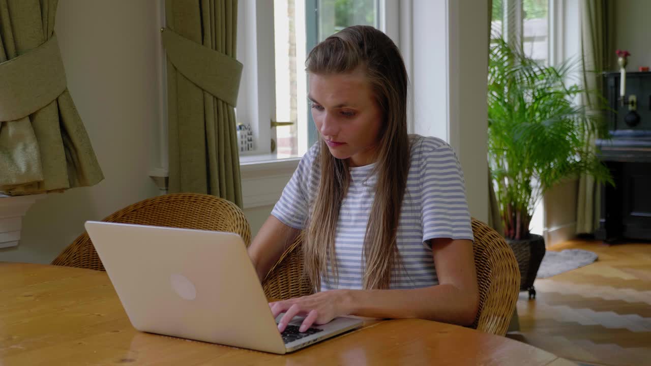 Young woman types on laptop on table at home, looks out window, slow motion