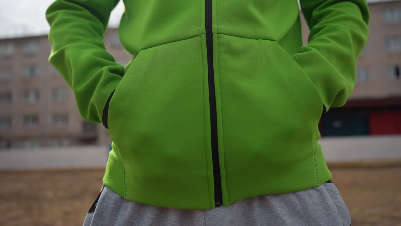 persona con un estilo de ropa relajado, modelos individuales con ropa casual en el exterior con las manos en los bolsillos, persona con chaqueta verde y pantalones de chándal grises de pie en el exterior con una postura relajada