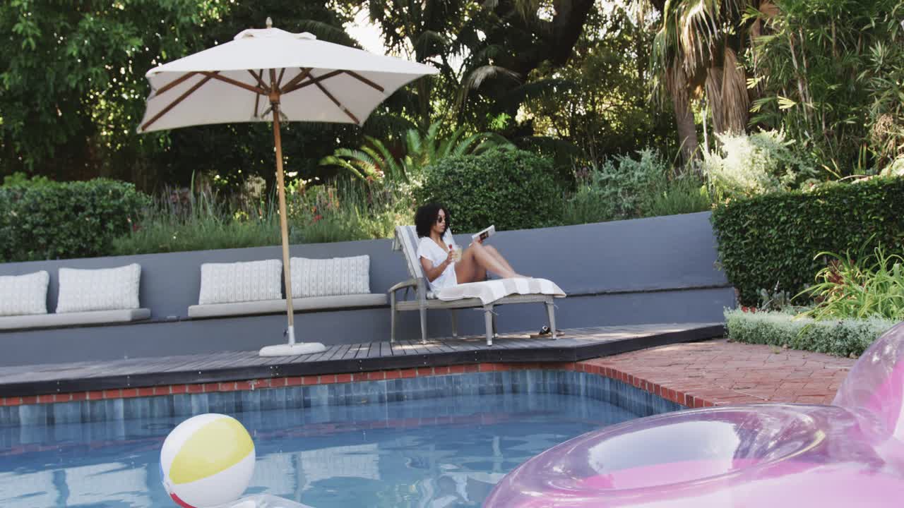 African American woman picking up book, reading on deck by pool with pink ring to relax