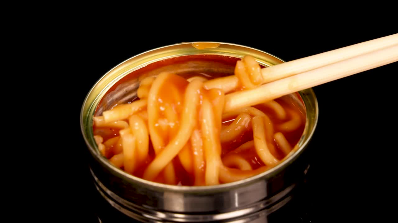Chopsticks grasp and lift spaghetti noodles in tomato sauce from a small metal can, shot against a black background with bright, even lighting