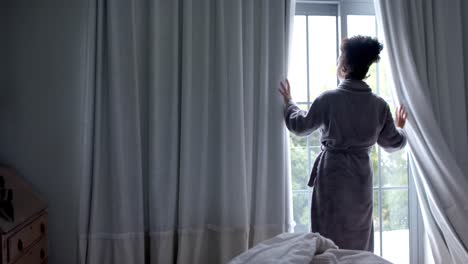 Back-of-biracial-woman-in-bathrobe-opening-curtains-in-bedroom,-slow-motion,-copy-space
