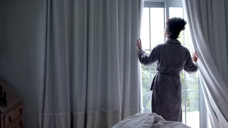 back of biracial woman in bathrobe opening curtains in bedroom, slow motion, copy space