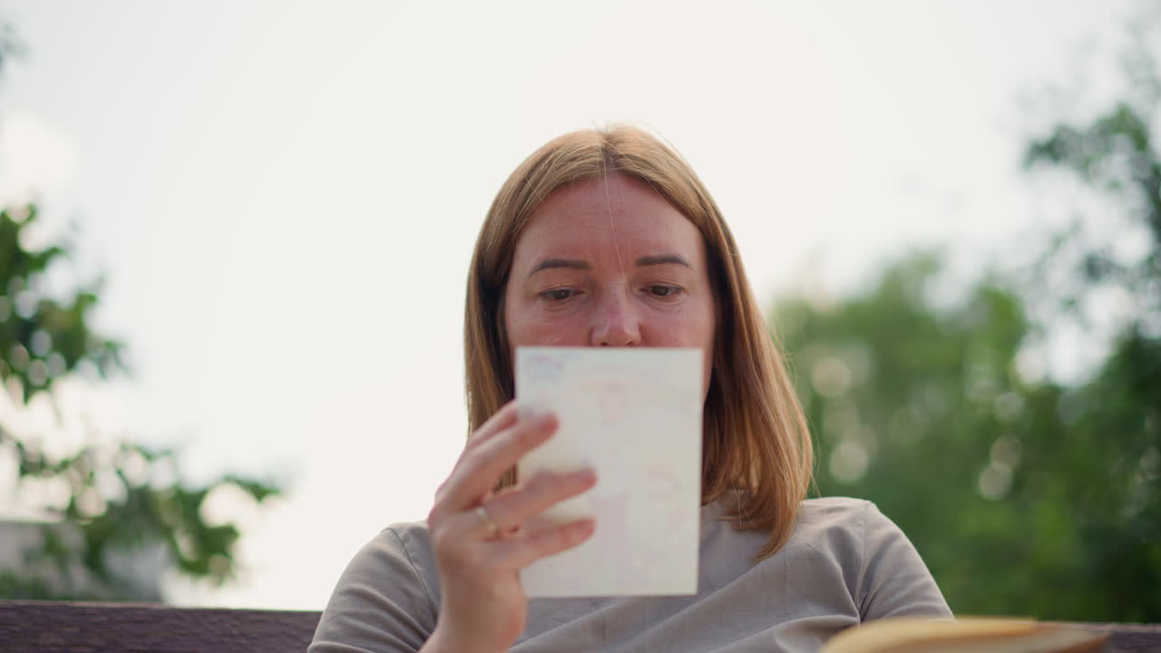 Close up portrait woman sitting outdoors holding old photo taken from book, gazing thoughtfully with calm expression under soft daylight, peaceful atmosphere blending nostalgia