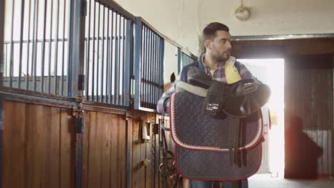 man is stroking a horse in stable while holding a saddle.