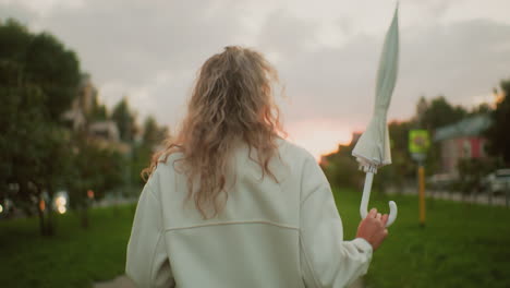 young adult wearing white coat walking along paved path twirling umbrella playfully during soft sunset with blurred view of signpost, trees, cars, and buildings
