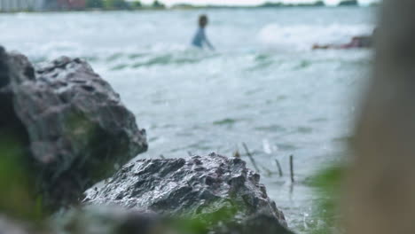 waves crash against rocks as surfers head out to the water in the background