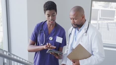 African-american-male-and-female-doctors-holding-clipboard-and-tablet,-talking-at-hospital