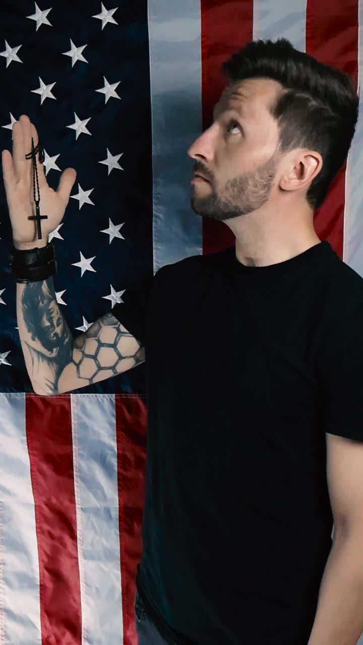 Tattooed man holding a black cross on a chain in his hands and looks up and start praying, religious man in front of an American vertical flag in slow motion. Looks up to god with faith and hope.