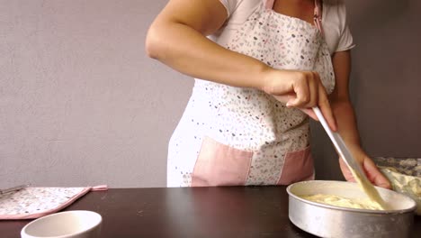 Mujer-Latina-Con-Un-Delantal-Preparando-La-Cocina-Horneando-Un-Pastel-Vertiendo-Y-Esparciendo-El-Doug-Con-Un-Raspador