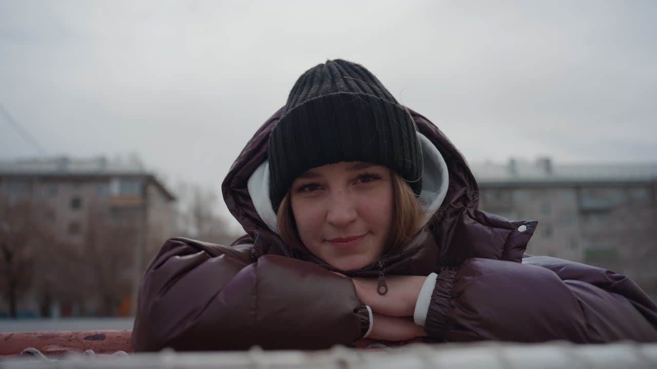 울타리에 기대어 야외에서 기다리는 흰 피부의 십 대, 갈색 패딩 점퍼와 니트 모자, 도시 옥상 및 아파트 배경, 흐린 하늘, 차분한 표정, 시네마틱한 차가운 톤과 아늑한 레이어링