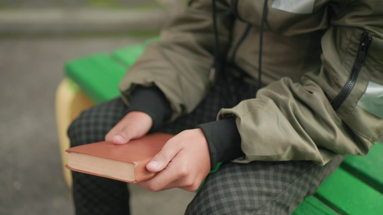 Close up partial view of person in checkered pants sitting on green bench tapping brown cover novel in left hand with subtle movement suggesting thoughtful gesture