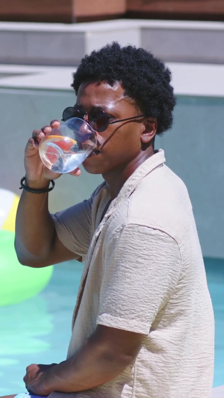 Vertical video: Sipping Black man in shades cooling from sun at pool holding cup with lemon, ring