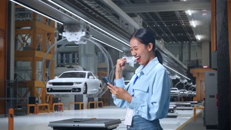 vista lateral de una trabajadora profesional asiática de pie con su teléfono inteligente en una fábrica de automóviles ev, instalación industrial de alta tecnología, levanta el puño con una meta gritando después de comprobar el teléfono inteligente