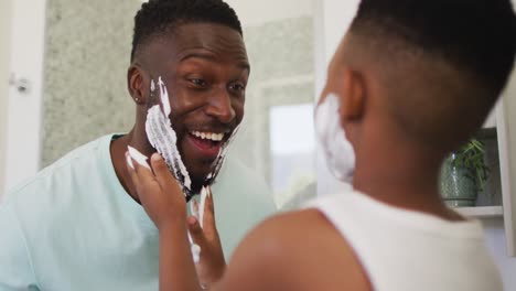 niño afroamericano poniendo crema de afeitar en la cara de su padre y riendo juntos