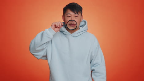 asian man holding magnifier glass on teeth, looking at camera showing funny smiling face mouth