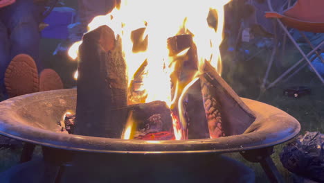 close up static shot of a camp fire, people sitting round the fire, large logs burning bright