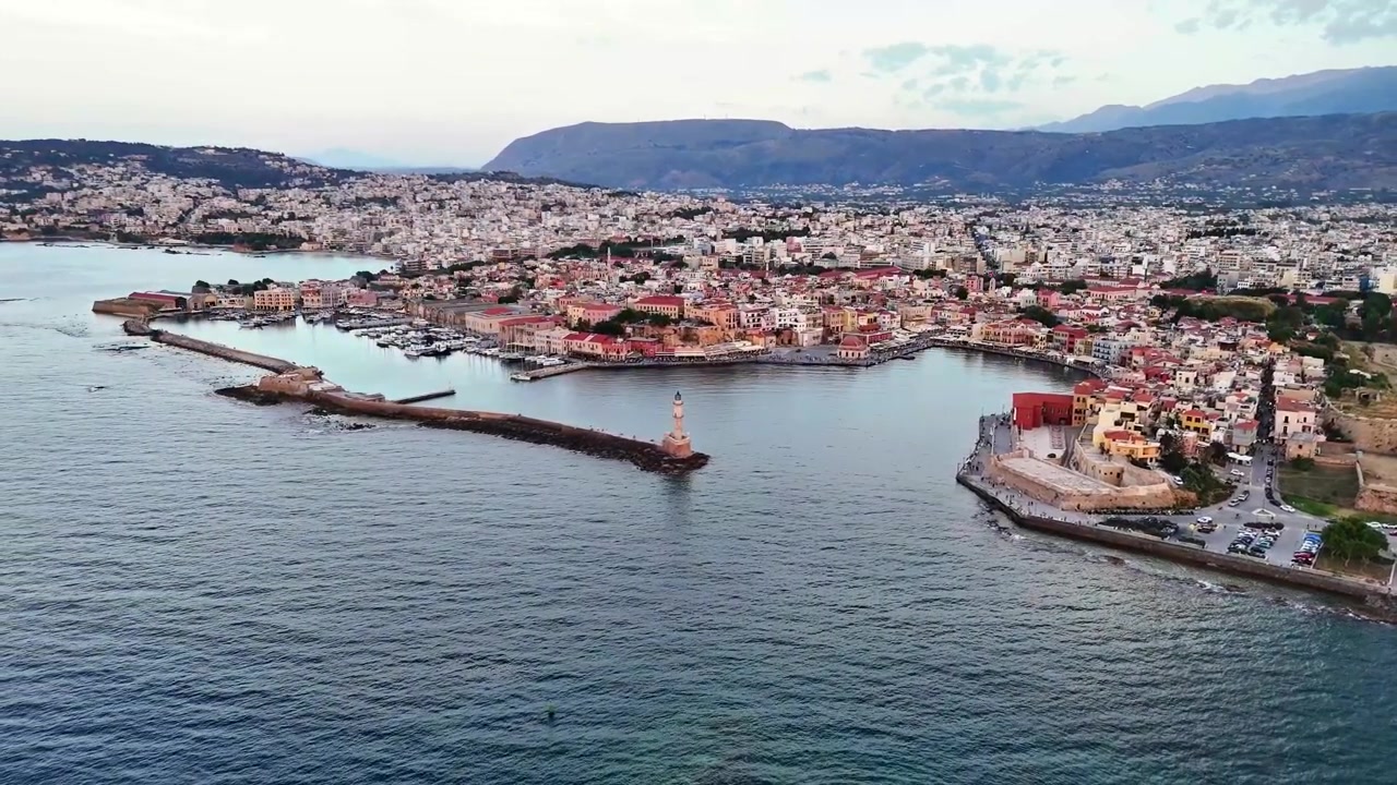 Yumuşak gün ışığı altında, uzun bir deniz feneri mendireği, tarihi sahil şeridi, yoğun şehir blokları ve dağ sıralarıyla birlikte Hanya limanı havadan çekim yapan kameranın altında beliriyor.