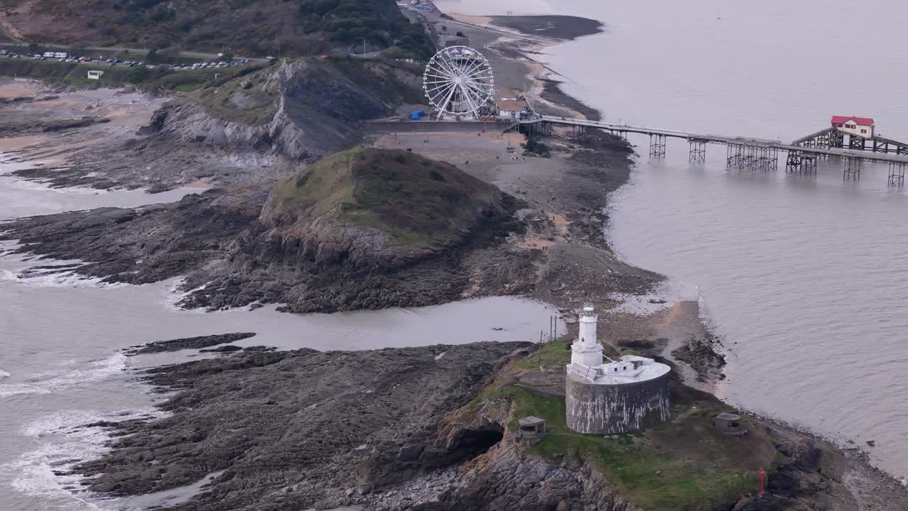 Drone telephoto orbit of Mumbles Pier in Swansea, showing the Big Wheel and waves rolling against the shore
