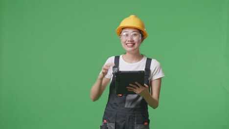 asian woman worker wearing goggles and safety helmet smiling and pointing to a tablet in her hand while standing in the green screen background studio