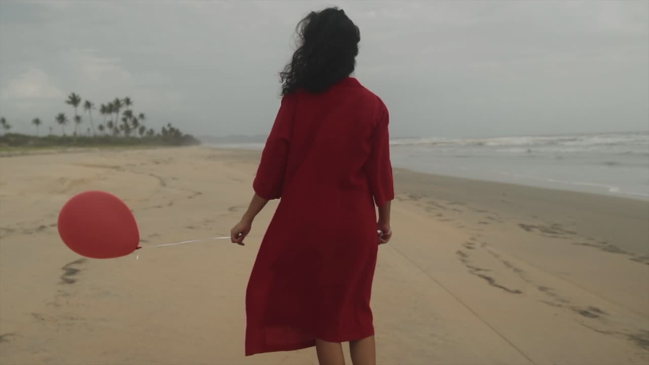 A woman strolling along the shoreline, adorned in a scarlet ensemble and gently holding a crimson balloon as she delightfully embraces the gentle caress of the breeze