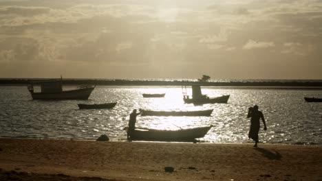 silhouette of two men preparing a small rowing boat to go fishing with a fishing cast net at a small bay full of anchored boats in the golden hour