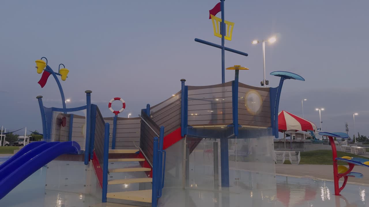 Camera moves slowly toward vibrant water play structure at empty pool, captured in slow motion