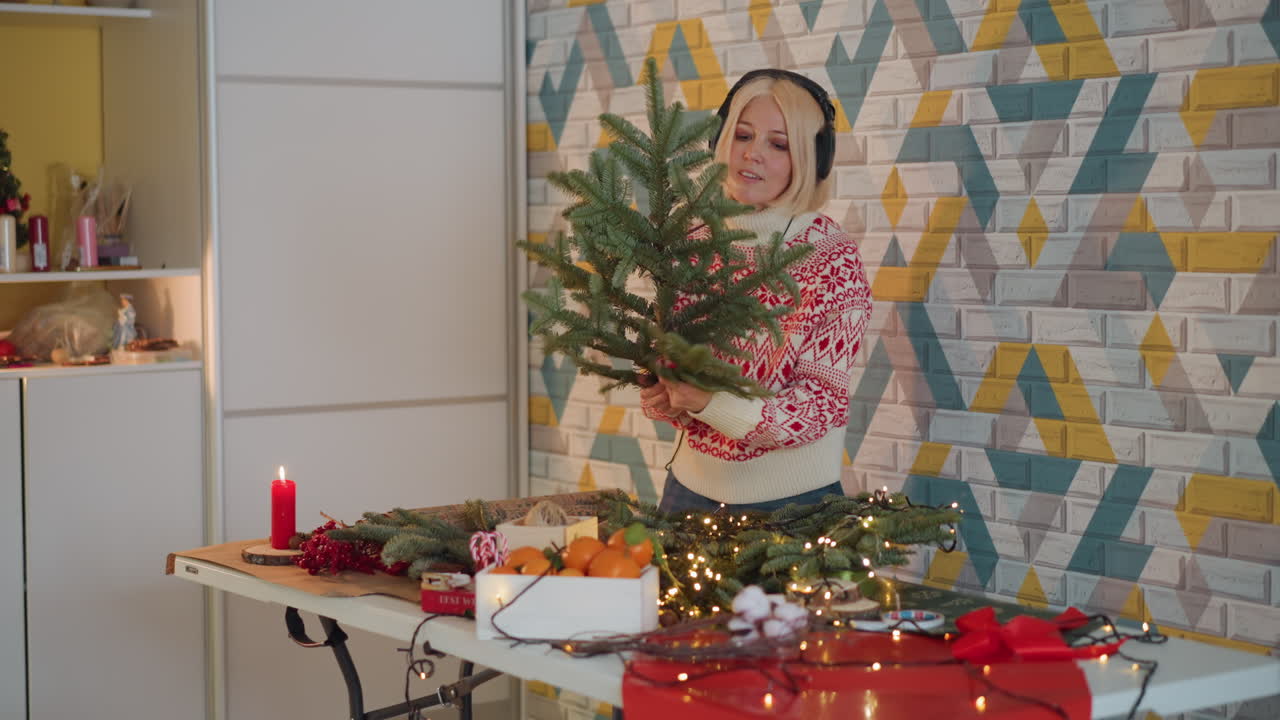 Mujer blanca creando una corona de Navidad en una mesa. Florista casera arreglando ramas de hoja perenne, canela, naranja, luz de velas y cinta en el espacio de trabajo de la cocina, vistiendo un suéter festivo y auriculares, ambiente lento y acogedor.