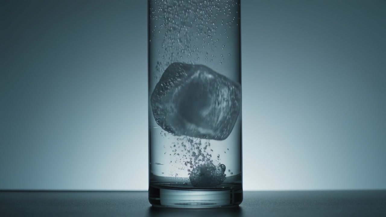 Una pastiglia effervescente che scintilla in un bicchiere alto sollevando un cubetto di ghiaccio su un tavolo da studio, le bollicine lo portano verso l'alto