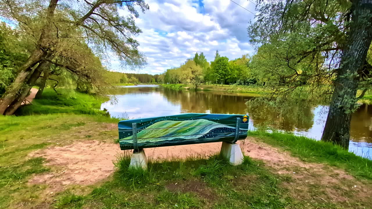 Painted bench with Camino de Santiago symbol beside Gauja river under cloudy sky