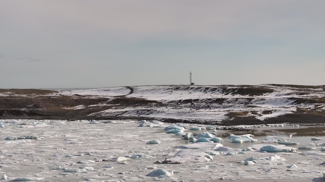 Icy chunks float on the still waters of Fjallsárlón, with snow-covered hills and a lone antenna tower rising on the horizon under a pale sky.