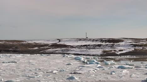 icy chunks float on the still waters of fjallsárlón, with snow-covered hills and a lone antenna tower rising on the horizon under a pale sky.