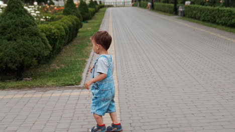 rear view of a little boy in jeans romper running by the paved road. child turns to camera, smiles and runs away.