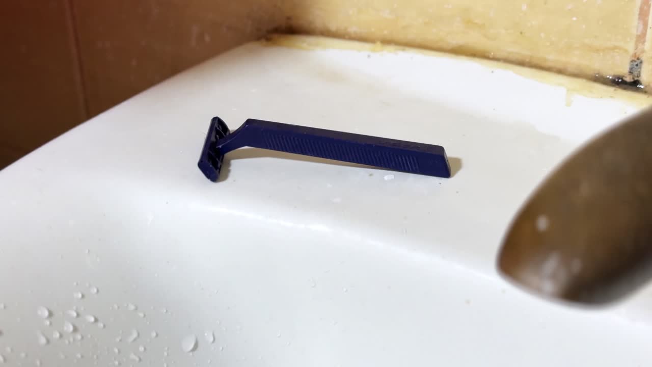 Close-up of a blue disposable razor resting on a white bathroom sink near a metal faucet, with visible water droplets and worn surfaces in an indoor hygiene setting