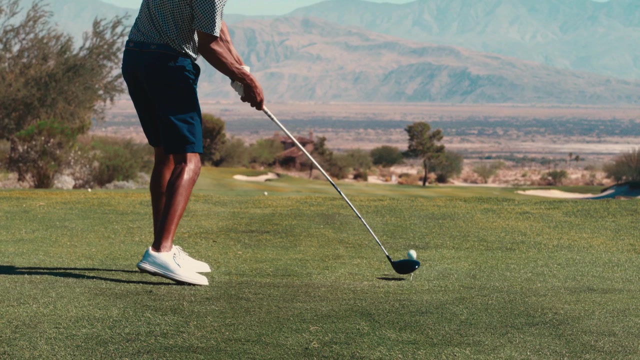 Un colpo di partenza con driver ripreso dal basso su un campo da golf nel deserto verso montagne distanti al rallentatore, adatto per spot di marchi di golf e contenuti di allenamento sulla meccanica dello swing