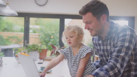 Padre-Trabajador-Usando-Una-Computadora-Portátil-En-Casa-En-El-Mostrador-De-La-Cocina-Mientras-Cuida-A-Su-Hija-Pequeña---Filmado-En-Cámara-Lenta