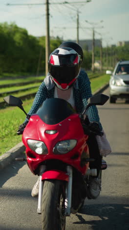 dos amigos montan una bicicleta eléctrica roja lentamente con cascos, cerca de una vía ferroviaria, un coche sigue detrás de ellos, y árboles verdes exuberantes se alinean en el lado opuesto de la carretera