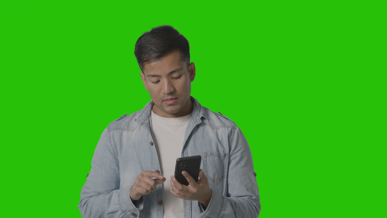 Studio-opname van casual geklede jongeman die online swipet op zijn mobiele telefoon tegen een groen scherm 2.