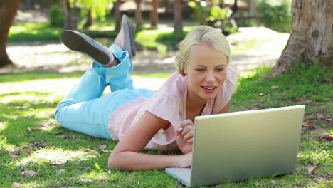 A-woman-lies-in-the-park-with-her-laptop-as-she-laughs-and-then-looks-at-the-camera